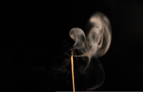 A delicate plume of smoke gracefully rises and curls against a dark background. The smoke emanates from a thin wooden incense stick, creating a sense of calm and tranquility.