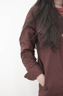 A person with long dark hair is wearing a maroon coat. The image is focused on the upper body, with one hand in the coat pocket. The background is plain and light-colored.