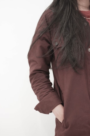 A person with long dark hair is wearing a maroon coat. The image is focused on the upper body, with one hand in the coat pocket. The background is plain and light-colored.