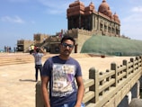 A man wearing sunglasses and a printed t-shirt stands in front of a historic stone monument with intricate architecture. The building has domes and pillars, and is set on a raised platform with stairs leading up to it. There are several tourists walking around and taking photos. The sky is clear with a few clouds, and the area is surrounded by a stone railing.