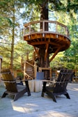 A wooden treehouse platform is nestled among tall trees, featuring a spiral staircase that wraps around the trunk. The structure is supported by sturdy beams and has a simple railing. Below, there is a wooden deck with several dark-colored Adirondack chairs arranged around a round table. The surrounding forest provides a serene and natural backdrop with sunlight filtering through the branches.