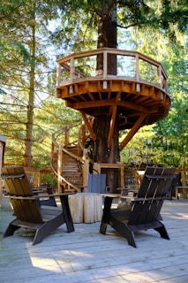 A wooden treehouse platform is nestled among tall trees, featuring a spiral staircase that wraps around the trunk. The structure is supported by sturdy beams and has a simple railing. Below, there is a wooden deck with several dark-colored Adirondack chairs arranged around a round table. The surrounding forest provides a serene and natural backdrop with sunlight filtering through the branches.