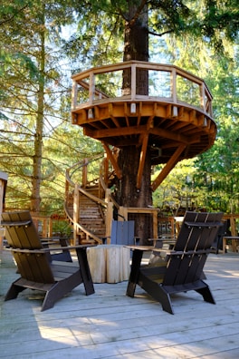 A wooden treehouse platform is nestled among tall trees, featuring a spiral staircase that wraps around the trunk. The structure is supported by sturdy beams and has a simple railing. Below, there is a wooden deck with several dark-colored Adirondack chairs arranged around a round table. The surrounding forest provides a serene and natural backdrop with sunlight filtering through the branches.