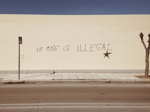 A large beige wall features graffiti with the words 'No One is Illegal' written in black, accompanied by a black star. In front of the wall, a narrow sidewalk and a street are visible, with a pedestrian sign on a pole and a leafless tree at the edge of the image. The scene is minimalistic and nearly deserted.