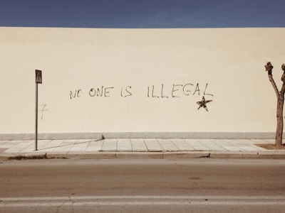 A large beige wall features graffiti with the words 'No One is Illegal' written in black, accompanied by a black star. In front of the wall, a narrow sidewalk and a street are visible, with a pedestrian sign on a pole and a leafless tree at the edge of the image. The scene is minimalistic and nearly deserted.