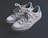 A pair of casual sneakers with white and gray tones are displayed against a dark background. The sneakers feature a prominent logo on the side, have a mesh and suede texture, with a classic lace-up design. The heel section is accented with a bright blue color.
