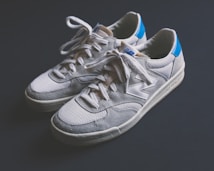 A pair of casual sneakers with white and gray tones are displayed against a dark background. The sneakers feature a prominent logo on the side, have a mesh and suede texture, with a classic lace-up design. The heel section is accented with a bright blue color.