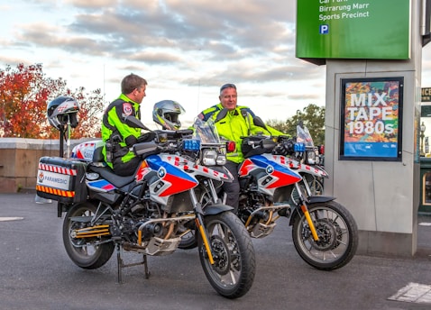Two paramedics in bright high-visibility jackets are sitting on motorcycles equipped for emergency medical services. The motorcycles are parked near a sign that reads 'MIX TAPE 1980s' and a parking information sign. The background features an autumn scene with trees displaying reddish leaves.