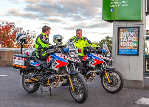 Two paramedics in bright high-visibility jackets are sitting on motorcycles equipped for emergency medical services. The motorcycles are parked near a sign that reads 'MIX TAPE 1980s' and a parking information sign. The background features an autumn scene with trees displaying reddish leaves.