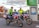 Two paramedics in bright high-visibility jackets are sitting on motorcycles equipped for emergency medical services. The motorcycles are parked near a sign that reads 'MIX TAPE 1980s' and a parking information sign. The background features an autumn scene with trees displaying reddish leaves.