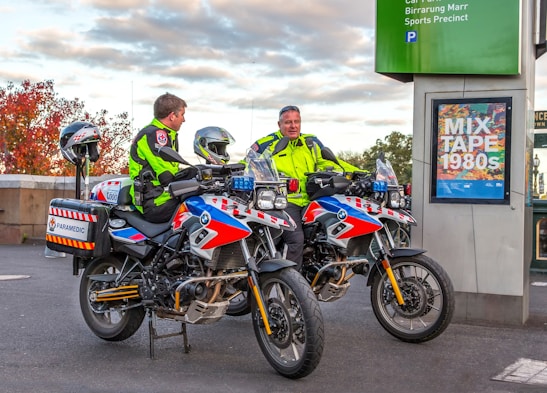 Two paramedics in bright high-visibility jackets are sitting on motorcycles equipped for emergency medical services. The motorcycles are parked near a sign that reads 'MIX TAPE 1980s' and a parking information sign. The background features an autumn scene with trees displaying reddish leaves.