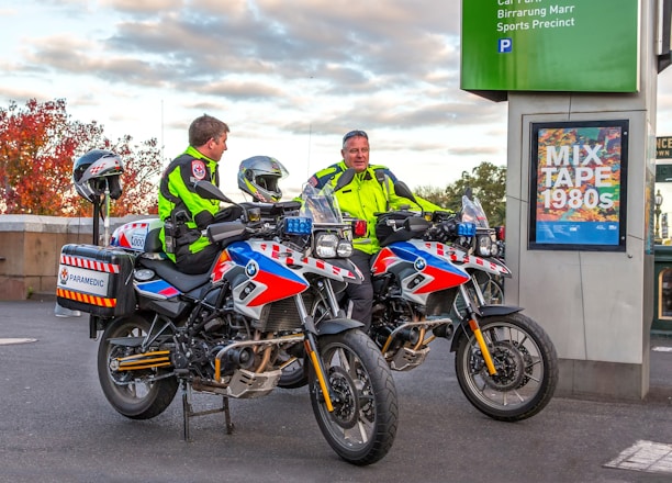 Two paramedics in bright high-visibility jackets are sitting on motorcycles equipped for emergency medical services. The motorcycles are parked near a sign that reads 'MIX TAPE 1980s' and a parking information sign. The background features an autumn scene with trees displaying reddish leaves.