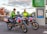 Two paramedics in bright high-visibility jackets are sitting on motorcycles equipped for emergency medical services. The motorcycles are parked near a sign that reads 'MIX TAPE 1980s' and a parking information sign. The background features an autumn scene with trees displaying reddish leaves.