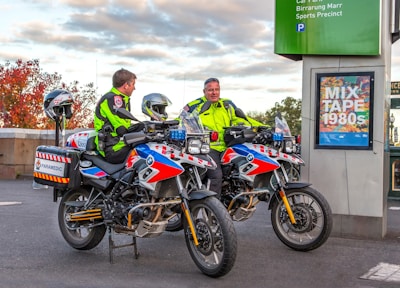 Two paramedics in bright high-visibility jackets are sitting on motorcycles equipped for emergency medical services. The motorcycles are parked near a sign that reads 'MIX TAPE 1980s' and a parking information sign. The background features an autumn scene with trees displaying reddish leaves.