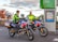 Two paramedics in bright high-visibility jackets are sitting on motorcycles equipped for emergency medical services. The motorcycles are parked near a sign that reads 'MIX TAPE 1980s' and a parking information sign. The background features an autumn scene with trees displaying reddish leaves.
