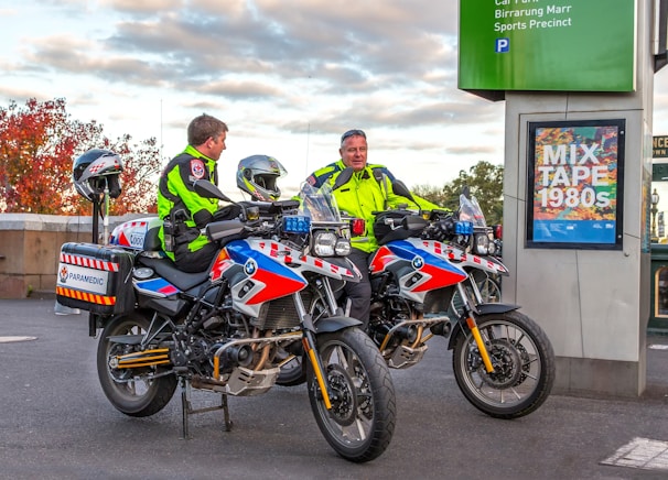 Two paramedics in bright high-visibility jackets are sitting on motorcycles equipped for emergency medical services. The motorcycles are parked near a sign that reads 'MIX TAPE 1980s' and a parking information sign. The background features an autumn scene with trees displaying reddish leaves.