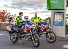 Two paramedics in bright high-visibility jackets are sitting on motorcycles equipped for emergency medical services. The motorcycles are parked near a sign that reads 'MIX TAPE 1980s' and a parking information sign. The background features an autumn scene with trees displaying reddish leaves.