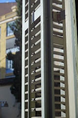 A close-up view of a window shutter with horizontal slats partially open, allowing light to filter through. The background features blurred elements of a building and a tree, indicating an urban outdoor scene.
