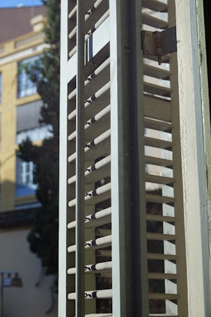 A close-up view of a window shutter with horizontal slats partially open, allowing light to filter through. The background features blurred elements of a building and a tree, indicating an urban outdoor scene.