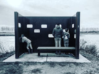 Three people are standing inside a dark, open shelter with rectangular openings. They appear to be looking through the wall toward a body of water. The surroundings include grassy and bushy vegetation and a tree-lined horizon.