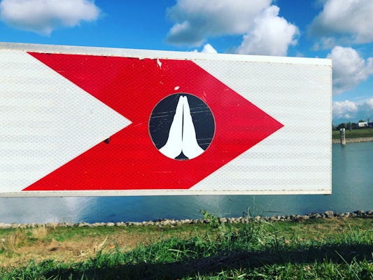 A sign with a large red arrow pointing to the right and a white icon of praying hands in the center is positioned against a backdrop of a grassy area and a body of water. The sky is clear with scattered white clouds.