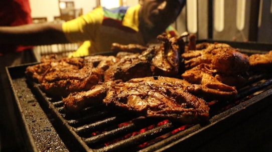 Several pieces of well-seasoned grilled chicken are sizzling on a barbecue grill. A person in the background appears to be tending to the grill, wearing a yellow and multicolored shirt. The scene is dimly lit, indicating it might be evening.