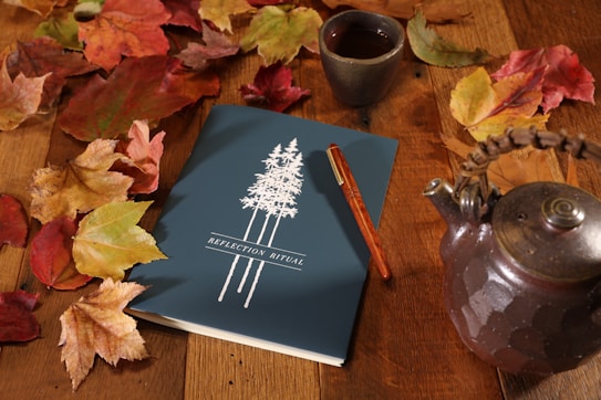 A blue notebook titled 'Reflection Ritual' is placed on a wooden surface surrounded by autumn leaves. A brown tea pot and a clay cup with liquid inside are nearby. A brown pen rests on the notebook, which has an illustration of tall trees on its cover.