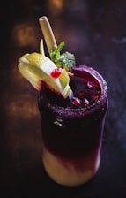 A dark, multicolored beverage in a glass is garnished with lime slices, pomegranate seeds, mint leaves, and served with a straw. The drink features a gradient from deep purple at the top to creamy at the bottom, with a sugared rim.