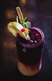 A dark, multicolored beverage in a glass is garnished with lime slices, pomegranate seeds, mint leaves, and served with a straw. The drink features a gradient from deep purple at the top to creamy at the bottom, with a sugared rim.