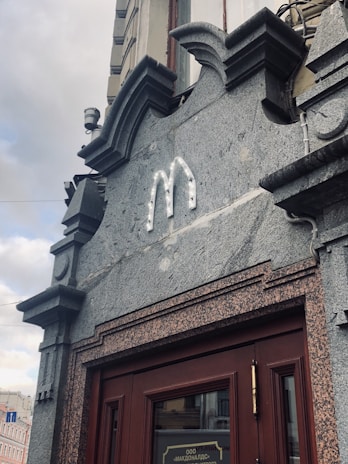 An ornate building facade features an unlit sign in the shape of an 'M'. The structure includes detailed architectural elements with stone and brick textures. Below the sign is a wooden door with reflective glass.