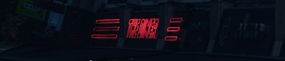 A series of neon signs are displayed on a building exterior, primarily featuring bold red lettering. The signs include text and geometric shapes, illuminated against a dark background, suggesting a nighttime urban setting.