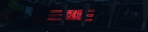 A series of neon signs are displayed on a building exterior, primarily featuring bold red lettering. The signs include text and geometric shapes, illuminated against a dark background, suggesting a nighttime urban setting.