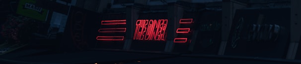 A series of neon signs are displayed on a building exterior, primarily featuring bold red lettering. The signs include text and geometric shapes, illuminated against a dark background, suggesting a nighttime urban setting.