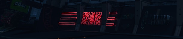 A series of neon signs are displayed on a building exterior, primarily featuring bold red lettering. The signs include text and geometric shapes, illuminated against a dark background, suggesting a nighttime urban setting.