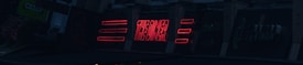 A series of neon signs are displayed on a building exterior, primarily featuring bold red lettering. The signs include text and geometric shapes, illuminated against a dark background, suggesting a nighttime urban setting.