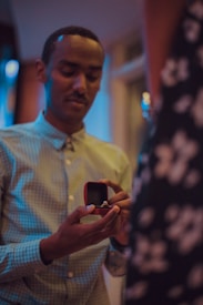 A person is holding an open ring box with a ring inside, likely proposing. The setting seems intimate with soft, warm lighting.