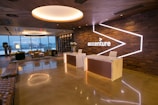 A modern office reception area with a polished floor and stylish wooden ceiling. The space features large windows offering a city view, contemporary seating arrangements, two reception desks with minimalist design, and a prominent accenture logo on an illuminated wall. There are also decorative elements such as potted plants and ambient lighting.