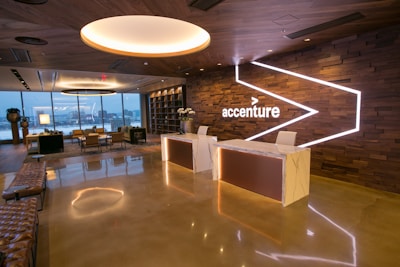 A modern office reception area with a polished floor and stylish wooden ceiling. The space features large windows offering a city view, contemporary seating arrangements, two reception desks with minimalist design, and a prominent accenture logo on an illuminated wall. There are also decorative elements such as potted plants and ambient lighting.