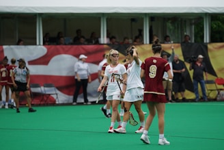 A group of female lacrosse players are on a green field. The players are wearing uniforms with one team in white and the other in maroon. They appear to be engaged in a discussion during a game. Spectators and coaches are visible in the background near the sidelines.