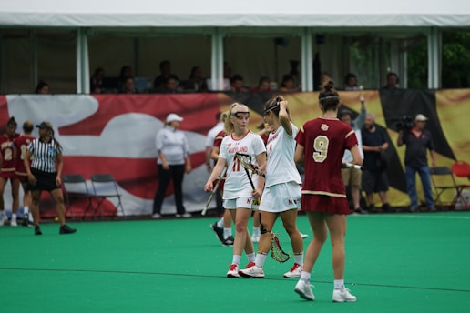 A group of female lacrosse players are on a green field. The players are wearing uniforms with one team in white and the other in maroon. They appear to be engaged in a discussion during a game. Spectators and coaches are visible in the background near the sidelines.