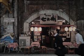 A small, cozy restaurant under a concrete archway displays traditional Japanese lanterns. Outside, there are tables with red checkered tablecloths and empty chairs. The facade is decorated with bottles and a wooden sign above the entrance. People in motion are blurred in the foreground, walking past the establishment.