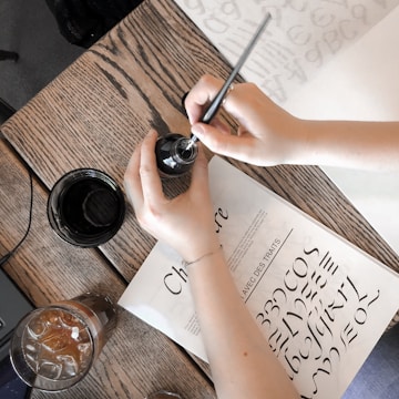 person holding bottle and pen over table with book and drinking glasses