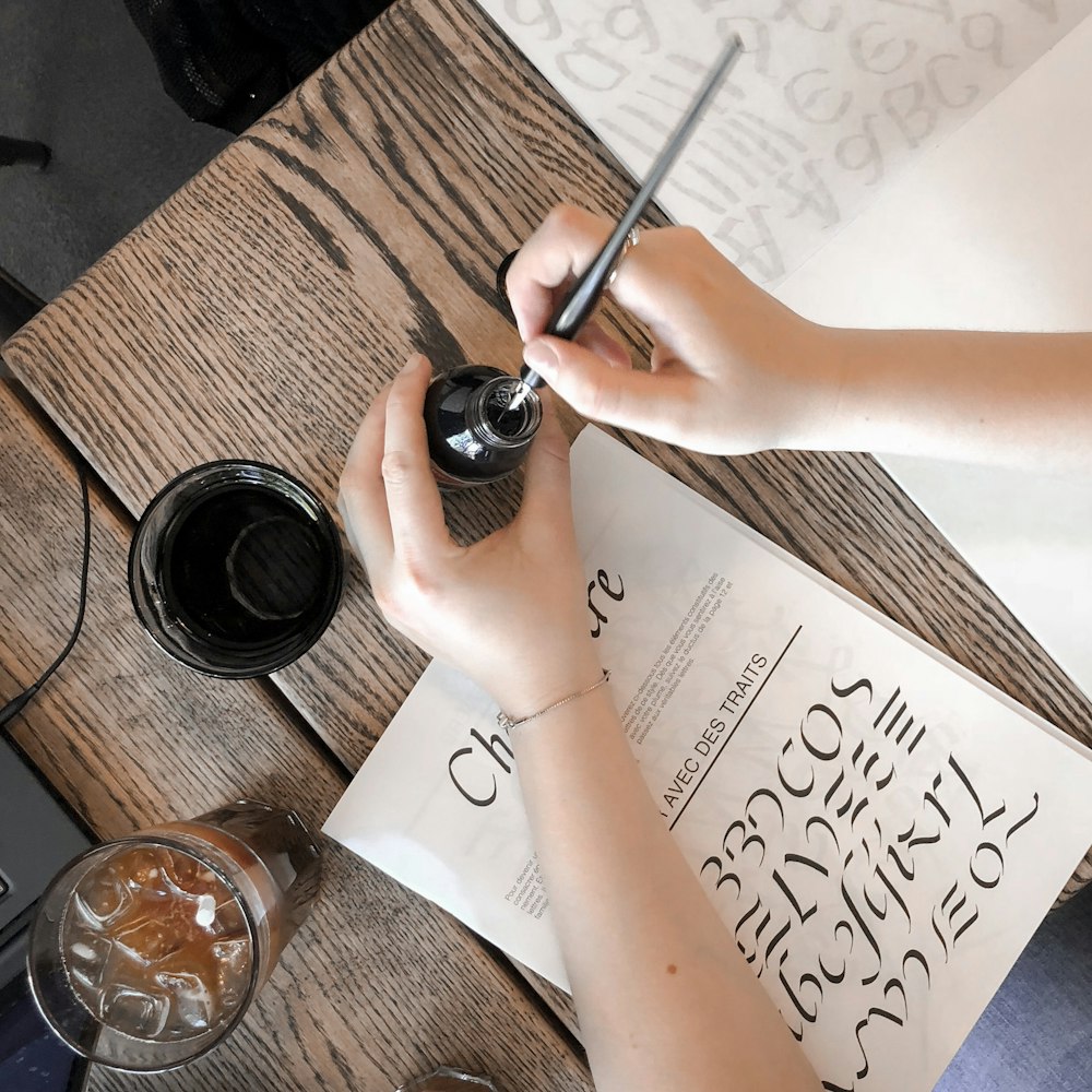 person holding bottle and pen over table with book and drinking glasses