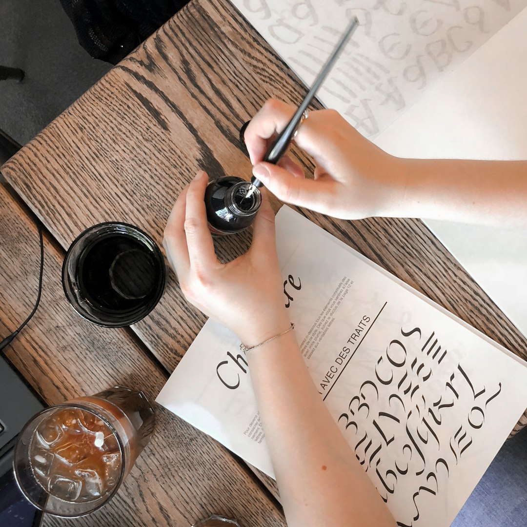 person holding bottle and pen over table with book and drinking glasses