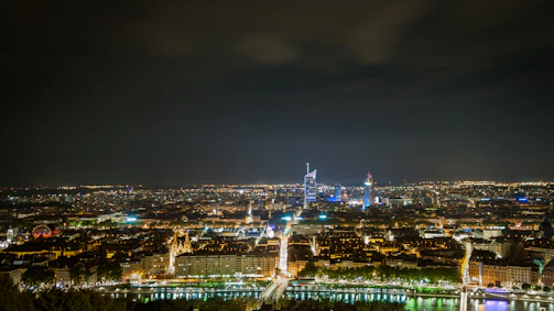 A bustling cityscape at night with glowing yellow lights reflecting on a river.
