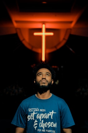 A man gazes upwards, standing below a prominently lit neon cross in a dimly lit environment. He wears a blue shirt with a spiritual quote printed on it.