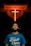 A man gazes upwards, standing below a prominently lit neon cross in a dimly lit environment. He wears a blue shirt with a spiritual quote printed on it.