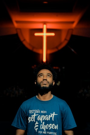 A man gazes upwards, standing below a prominently lit neon cross in a dimly lit environment. He wears a blue shirt with a spiritual quote printed on it.