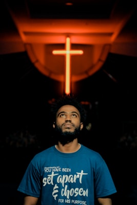 A man gazes upwards, standing below a prominently lit neon cross in a dimly lit environment. He wears a blue shirt with a spiritual quote printed on it.