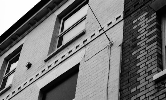 A monochrome view of a building's facade, featuring a mix of brick and tiled patterns. Two windows are visible, one is open slightly, and a security camera is mounted on the wall. There are wires running vertically across the surface.